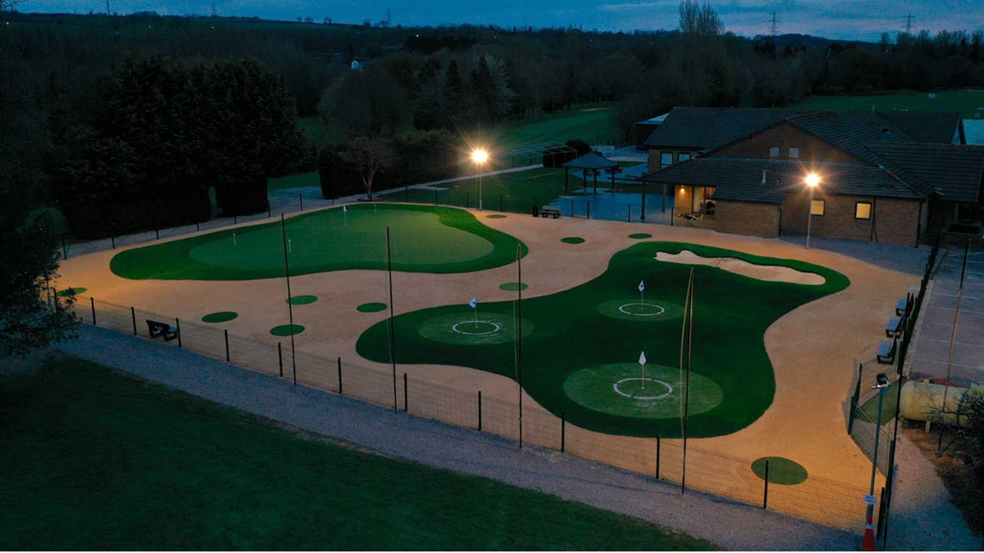 Aerial view of a large commercial putting green with multiple holes, sand bunkers, and chipping targets illuminated by floodlights at dusk