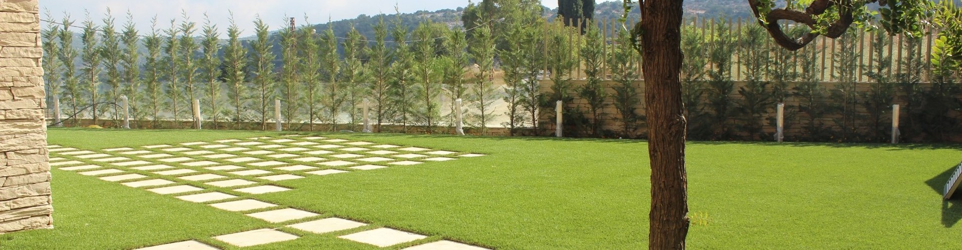 Southwest Greens lawn turf grass with a grid of square stepping stones, mature shade tree, and row of young cypress trees along a hillside wall.